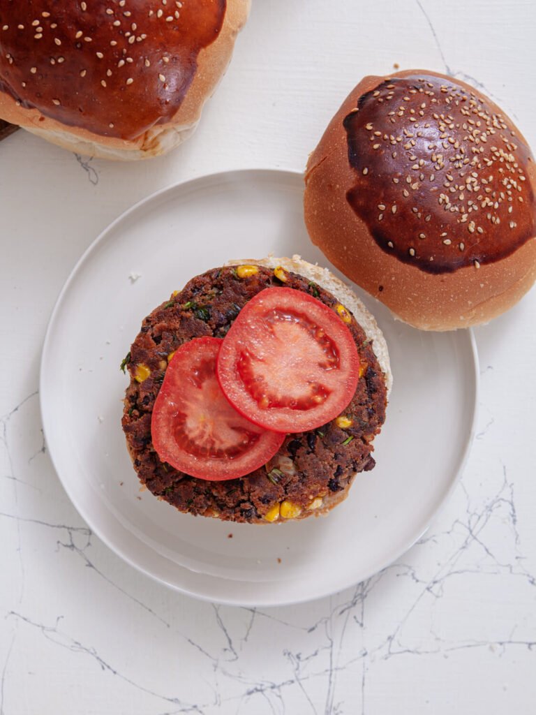 A black bean veggie burger on a white plate, topped with sliced tomatoes and ready to be assembled with the top sesame seed bun placed beside it
