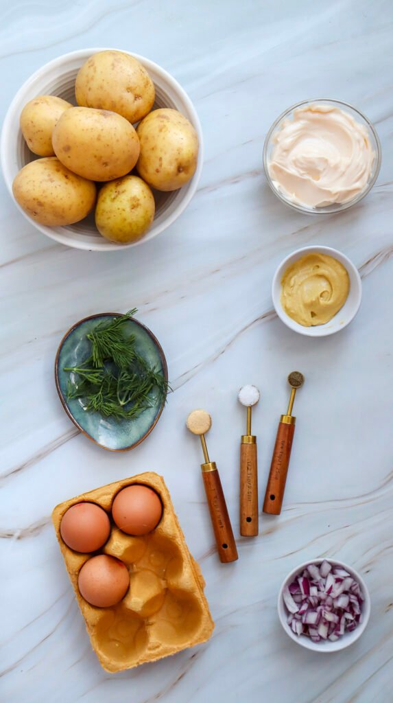 Ingredients for potato salad including potatoes, eggs, mayo, mustard, red onion, dill, and seasonings on a marble surface