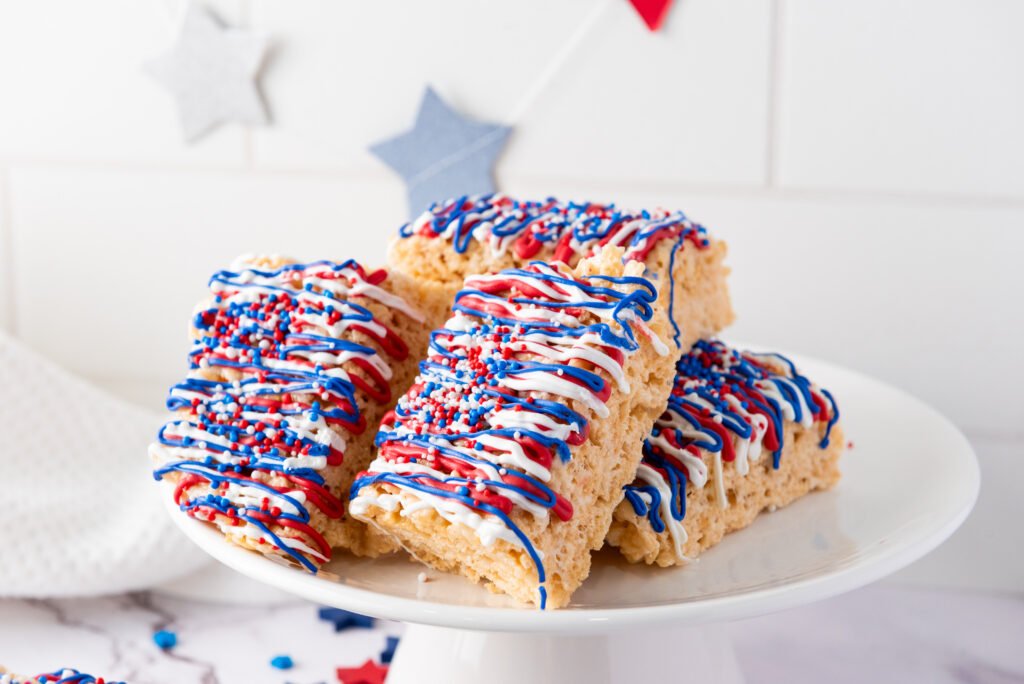 Stack of 4th of July Rice Krispie Treats topped with red, white, and blue drizzle and sprinkles, served on a white plate