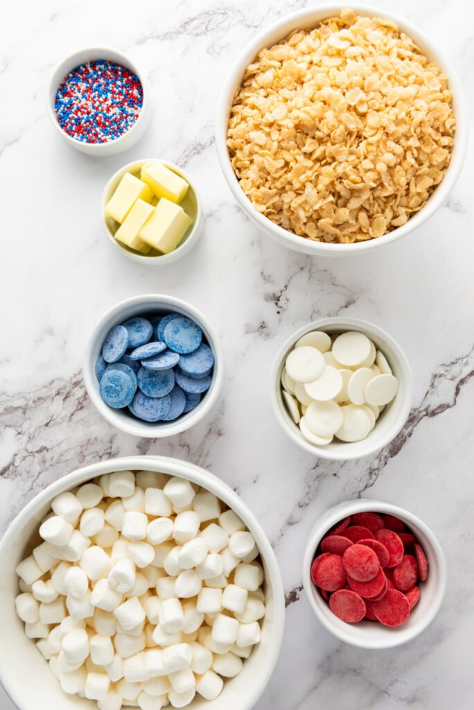 Ingredients for 4th of July Rice Krispie Treats laid out in bowls, including Rice Krispies cereal, mini marshmallows, butter, red, white, and blue candy melts, and patriotic sprinkles