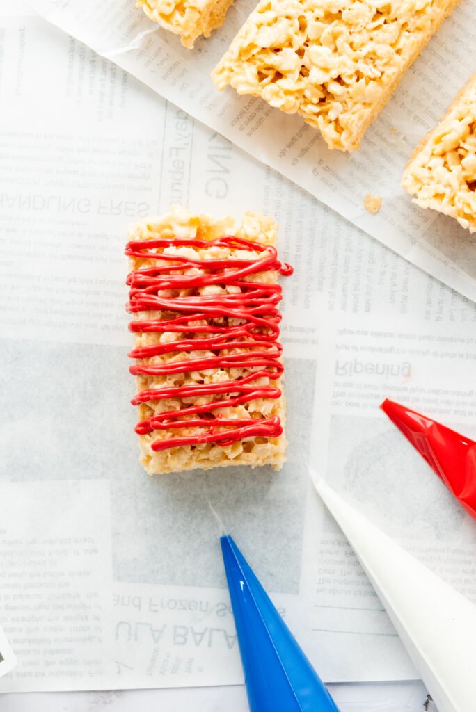 4th of July Rice Krispie treat bar drizzled with red candy melt, shown on parchment paper with blue and white piping bags nearby