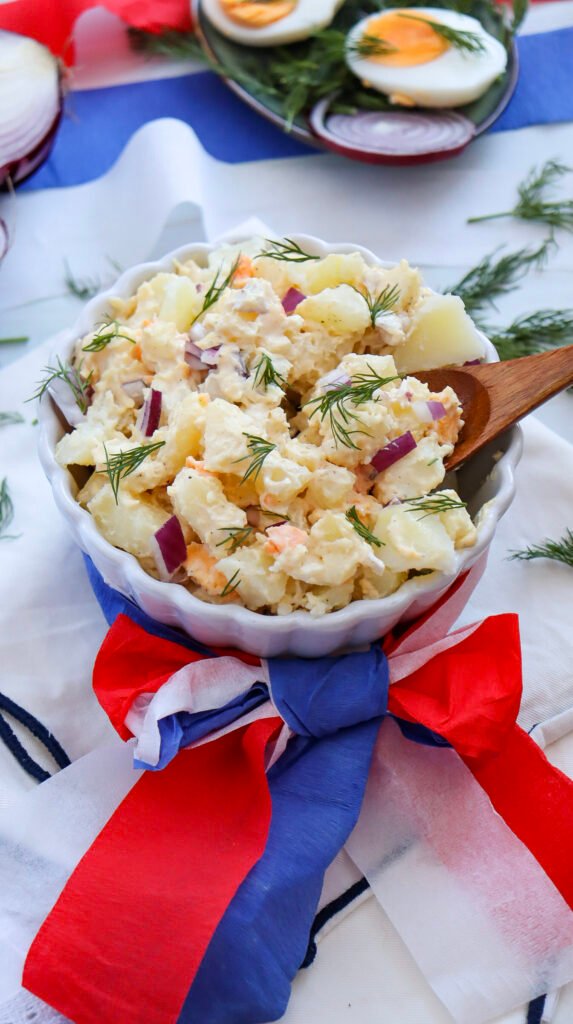 Bowl of creamy potato salad with red onions and dill, decorated with a red, white, and blue ribbon for July 4th