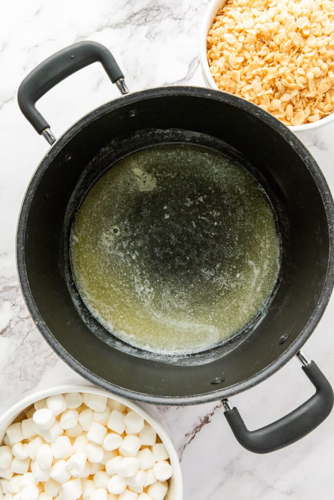 Large pot with melted butter inside, next to bowls of mini marshmallows and Rice Krispies cereal