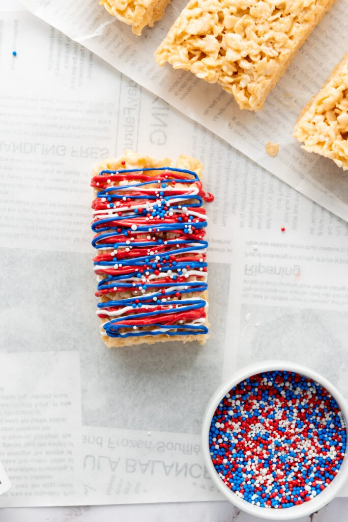 Decorated Rice Krispie treat with red, white, and blue drizzle and sprinkles, placed on parchment paper next to a small bowl of patriotic sprinkles