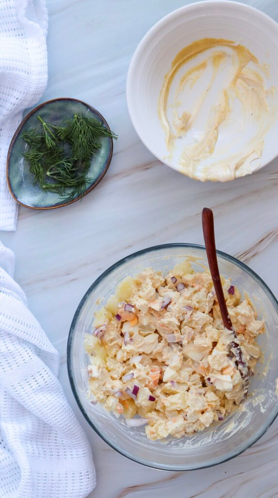 Mixed potato salad in a glass bowl with a wooden spoon, next to an empty dressing bowl and a plate of fresh dill