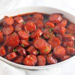 Sliced kielbasa baked in BBQ sauce, served in a white oval dish and garnished with chopped parsley
