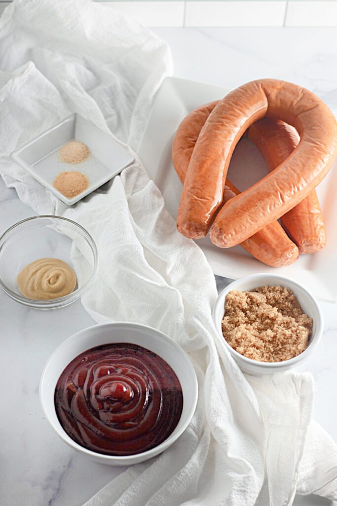 Ingredients for BBQ Baked Kielbasa arranged on a white surface, including whole kielbasa, BBQ sauce, brown sugar, Dijon mustard, and small bowls of garlic and onion powder