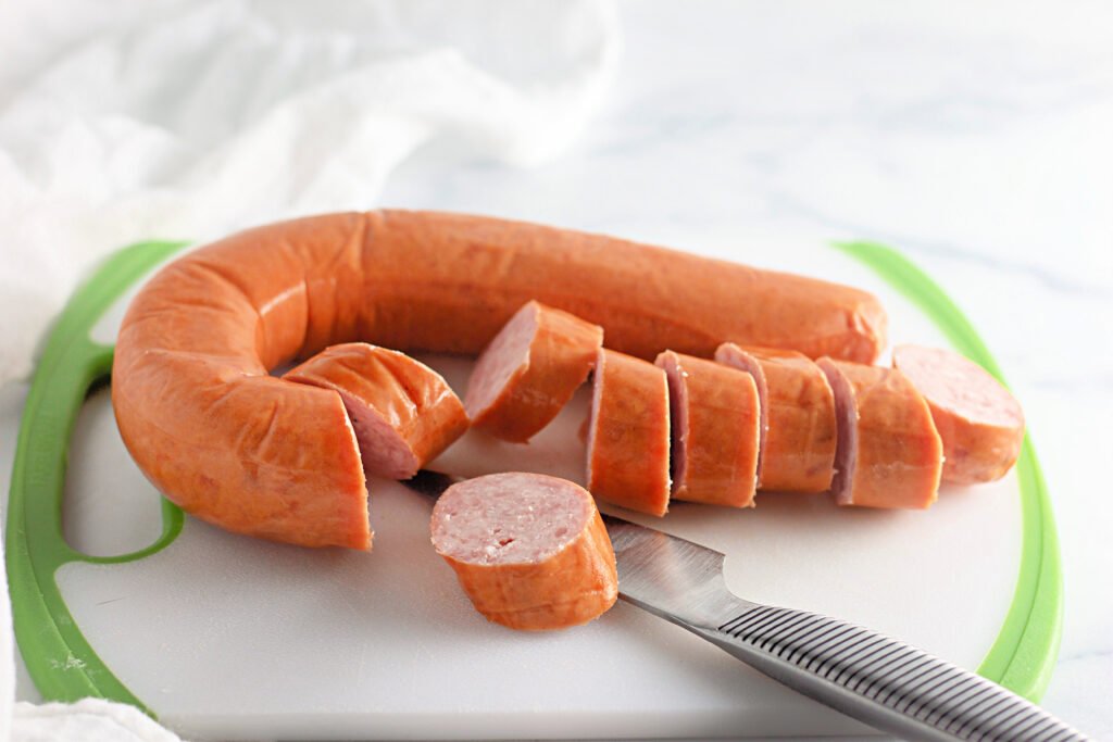 Kielbasa sausage being sliced on a white and green cutting board with a knife beside it