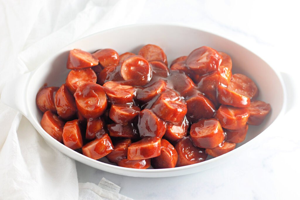 Sliced kielbasa coated in BBQ sauce, arranged in a white oval baking dish