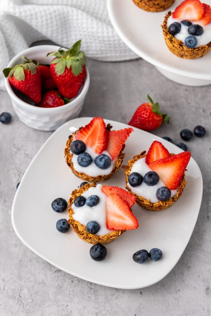 Granola cups topped with yogurt, strawberries, and blueberries on a white plate, with fresh berries scattered around