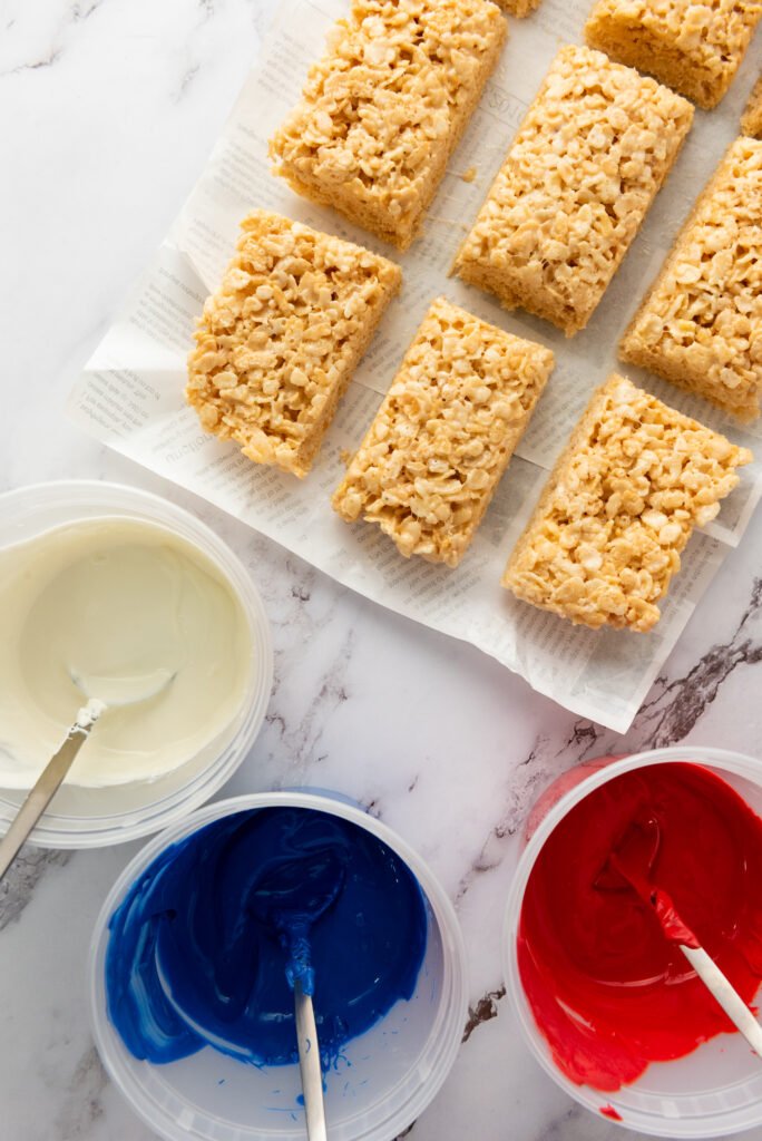 Rice Krispie treats arranged on parchment paper next to bowls of melted red, white, and blue candy melts