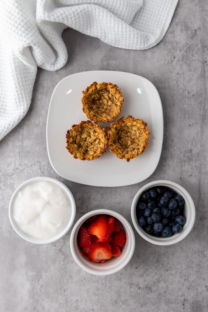 Baked granola cups on a plate with small bowls of yogurt, sliced strawberries, and blueberries for filling