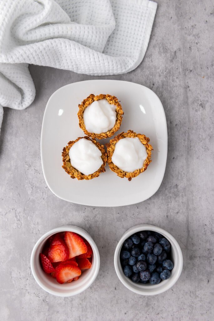 Granola cups filled with yogurt on a plate, with bowls of sliced strawberries and blueberries nearby