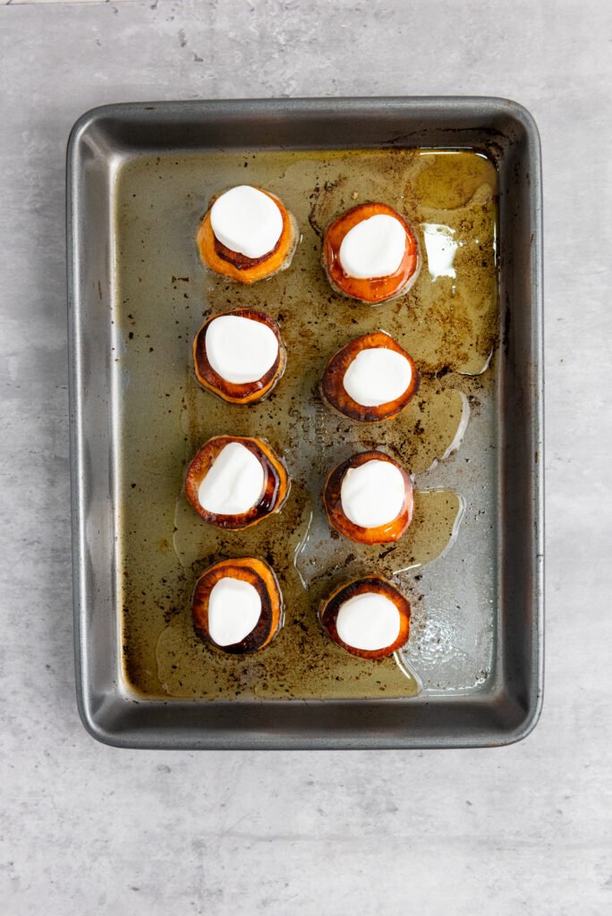 Roasted sweet potato slices on a baking tray, each topped with a halved marshmallow before broiling