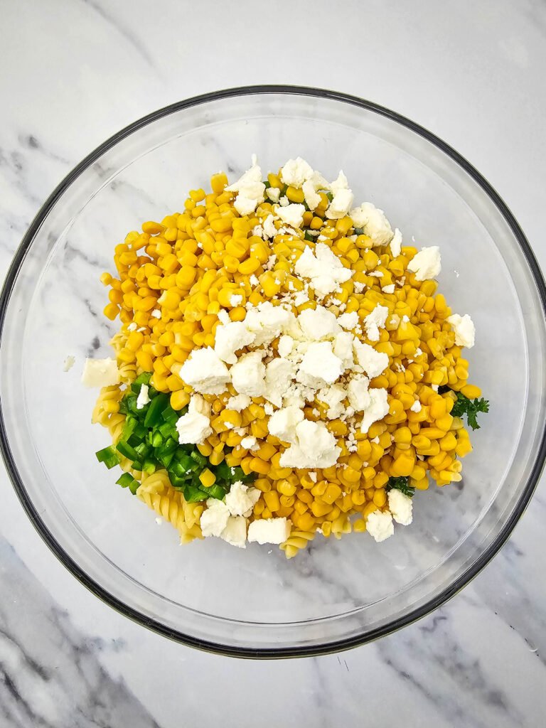 Unmixed ingredients for Mexican street corn pasta salad in a glass bowl, including corn, jalapeños, parsley, cotija cheese, and rotini pasta
