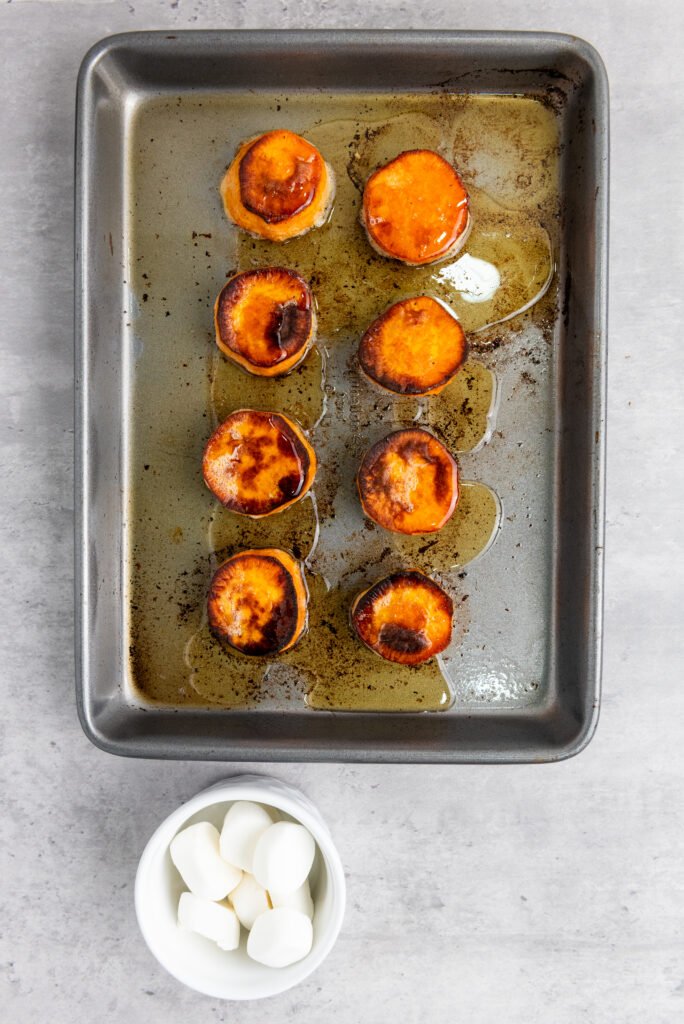 Roasted sweet potato slices on a baking tray with melted maple syrup, next to a small white bowl of marshmallows