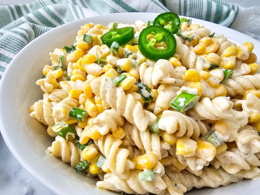 A close-up of creamy Mexican street corn pasta salad in a white bowl, made with rotini, corn, green onions, jalapeños, and cotija cheese