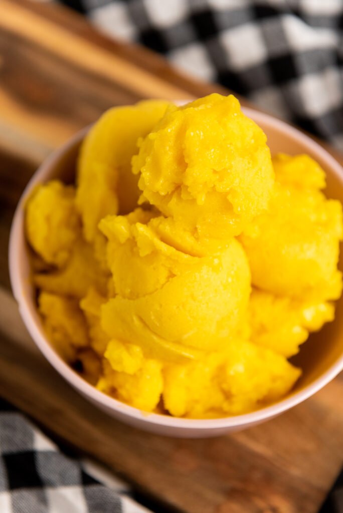 scoops of no-churn vegan mango sorbet in a pink bowl, placed on a wooden board with a checkered cloth in the background