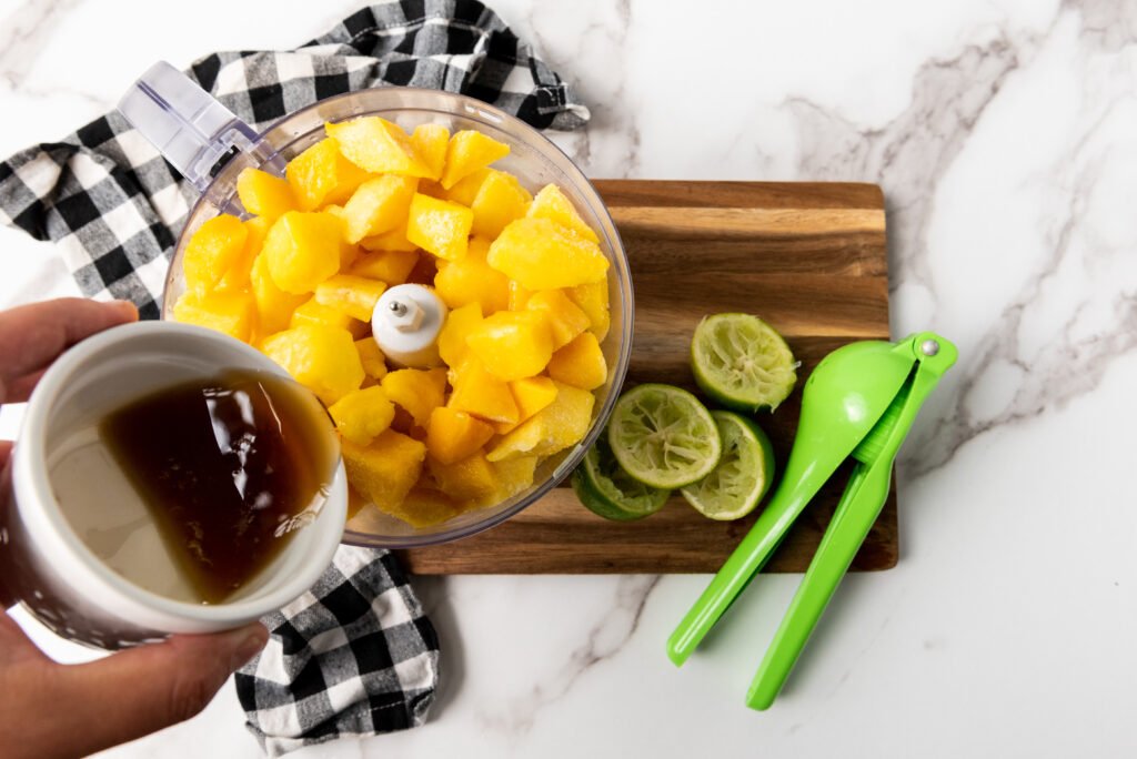 Maple syrup being poured into a food processor filled with frozen mango chunks, with squeezed lime halves and a green citrus squeezer on the side