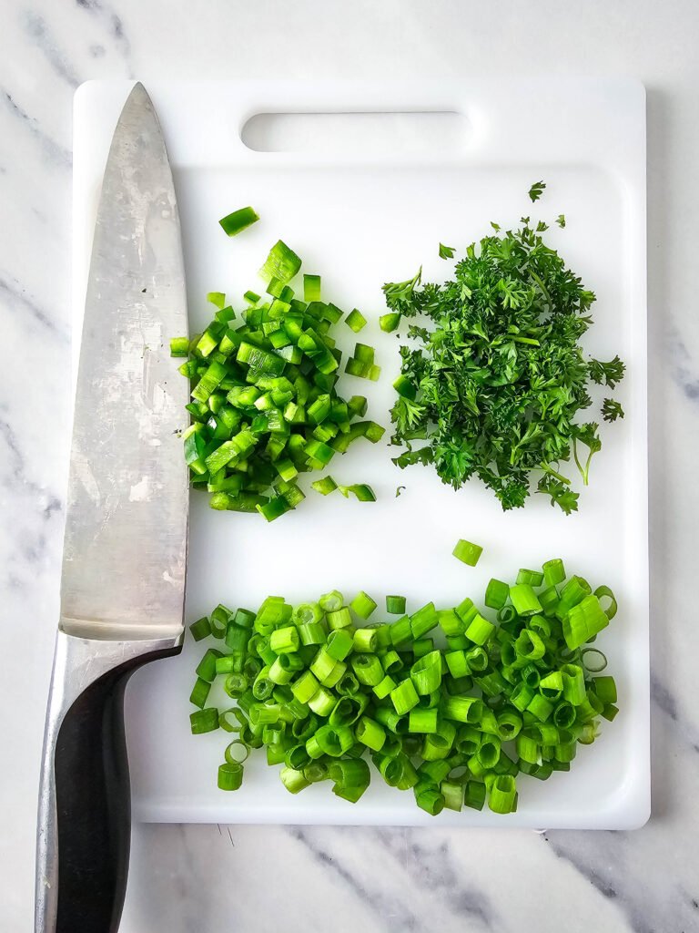 Chopped green onions, parsley, and jalapeño on a white cutting board with a kitchen knife, ready to be added to the Mexican street corn pasta salad