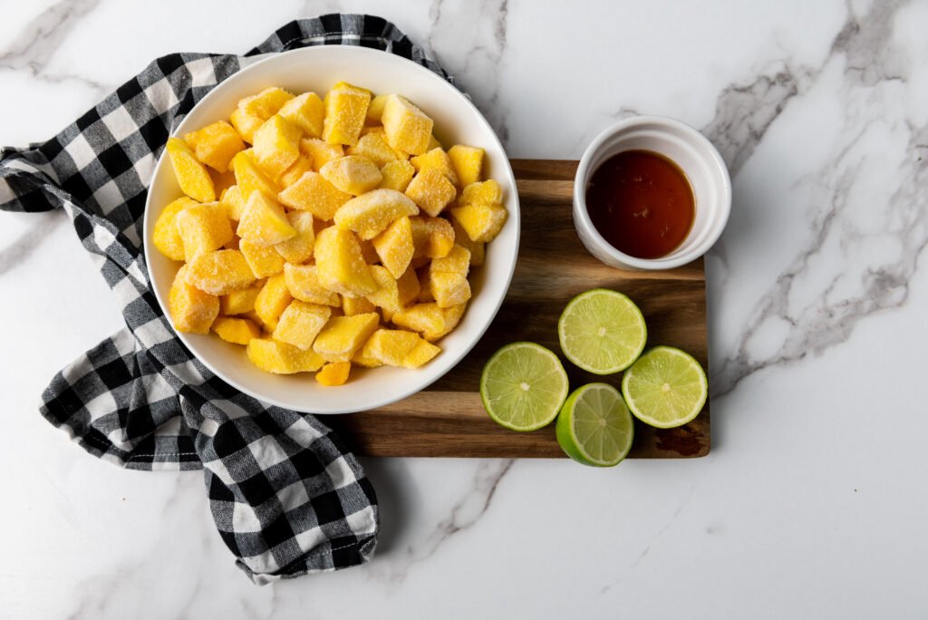 A bowl of frozen mango chunks next to a small ramekin of maple syrup and sliced limes on a wooden board