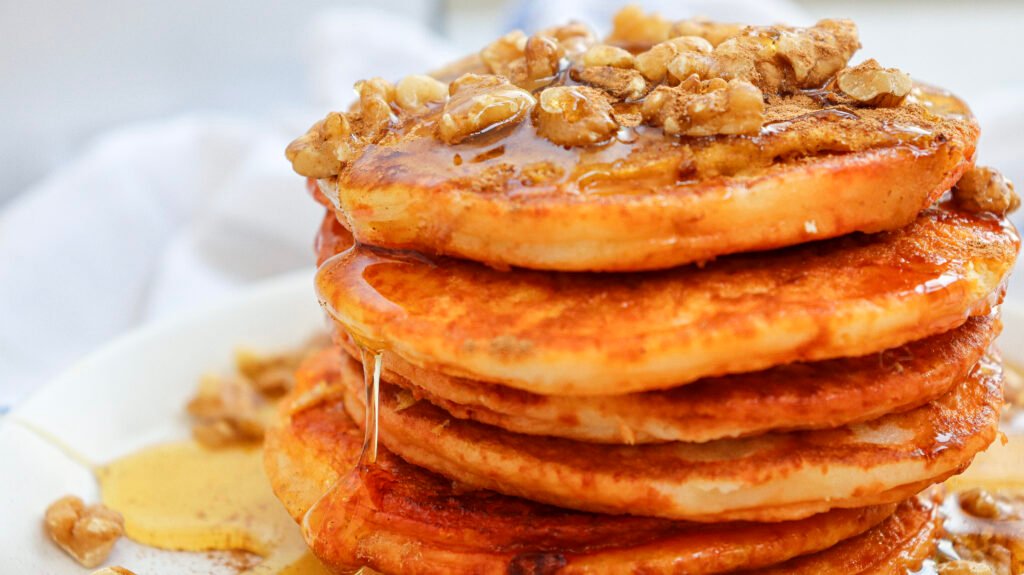 Stack of golden pumpkin pancakes topped with chopped walnuts and drizzled with maple syrup, with syrup dripping down the sides