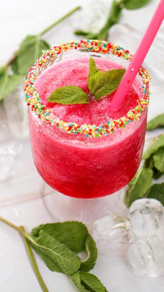 A vibrant pink watermelon slushie served in a clear glass with a colorful sprinkle-coated rim