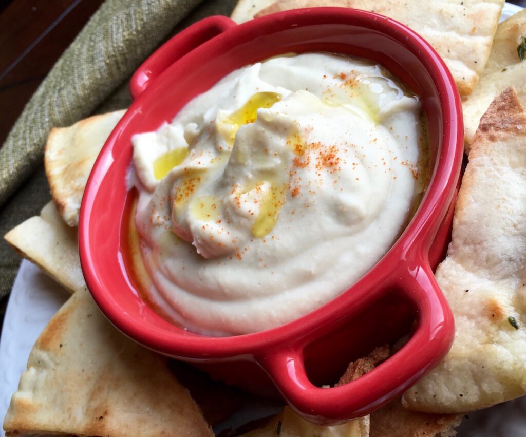 Creamy white bean dip in a red bowl, drizzled with olive oil and sprinkled with cayenne pepper