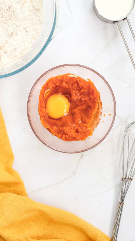 Pumpkin purée and a cracked egg in a glass bowl, placed next to a bowl of dry ingredients and a cup of milk on a white surface