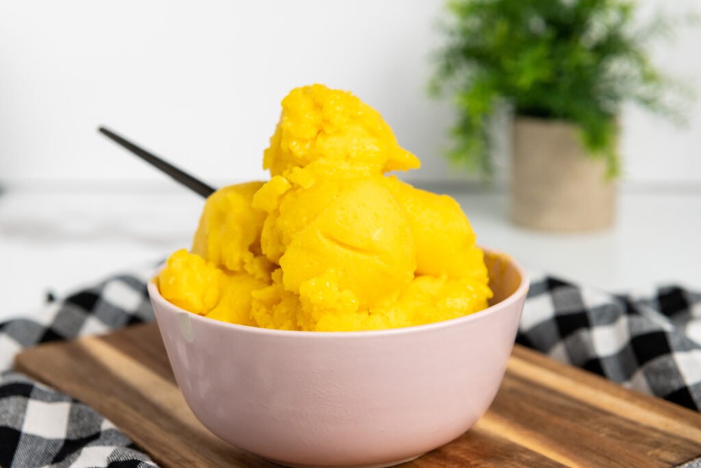 Scoops of homemade vegan mango sorbet in a light pink bowl, set on a wooden board with a checkered cloth and a green plant in the background