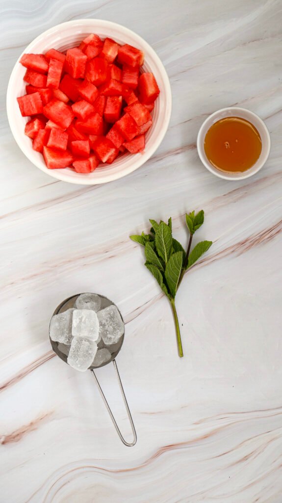 Ingredients for a homemade watermelon slushie arranged on a light marble surface, it shows cubed watermelon, honey, fresh mint leaves and ice cubes