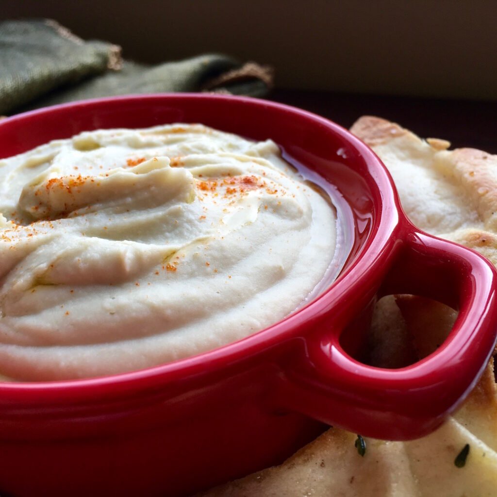 Creamy white bean dip in a red bowl, drizzled with olive oil and sprinkled with cayenne pepper