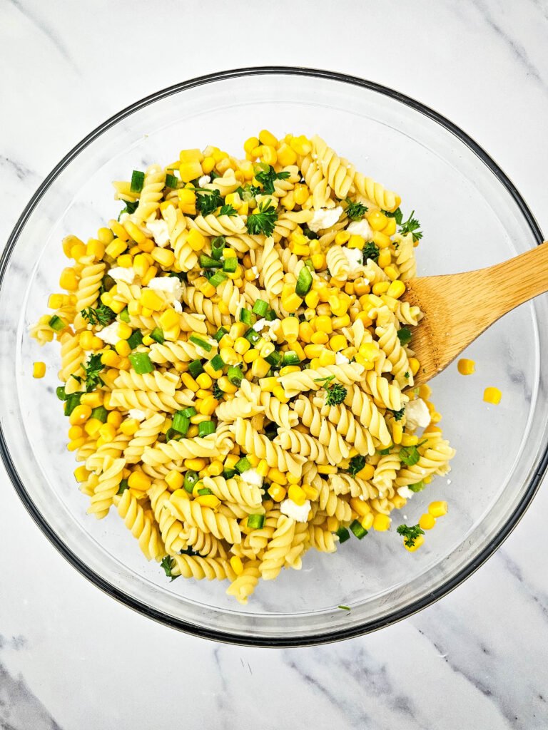 Mexican street corn pasta salad partially mixed in a glass bowl with a wooden spoon, showing corn, pasta, green onions, parsley, and cotija cheese
