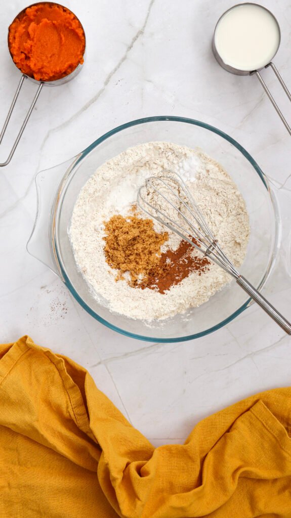 Dry ingredients for pumpkin pancakes in a glass bowl with a whisk, surrounded by pumpkin purée, milk, and a yellow kitchen towel