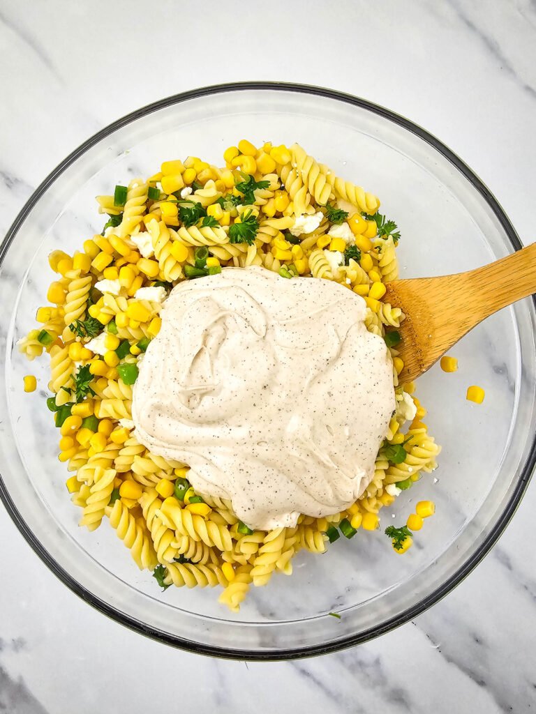 Creamy dressing placed on top of the corn, pasta, and chopped vegetables in a glass bowl, ready to be mixed into the Mexican street corn pasta salad