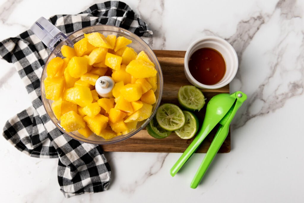 Frozen mango chunks in a food processor, surrounded by squeezed lime halves, a cup of maple syrup, and a green citrus press on a wooden board