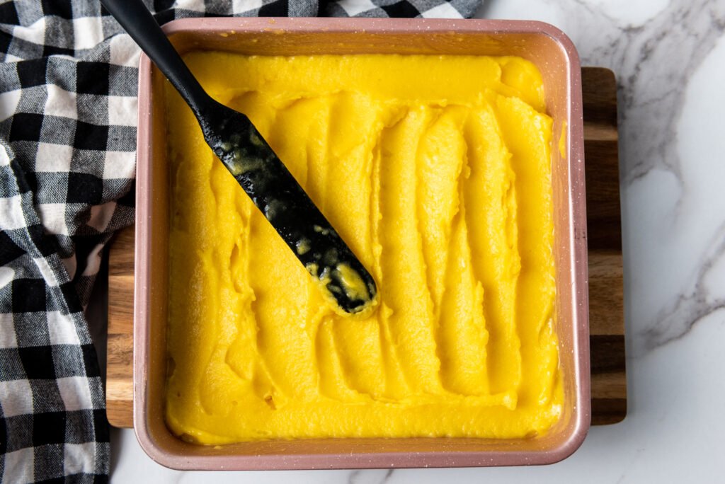 A baking dish filled with smooth mango sorbet being spread with a black spatula, ready to be frozen