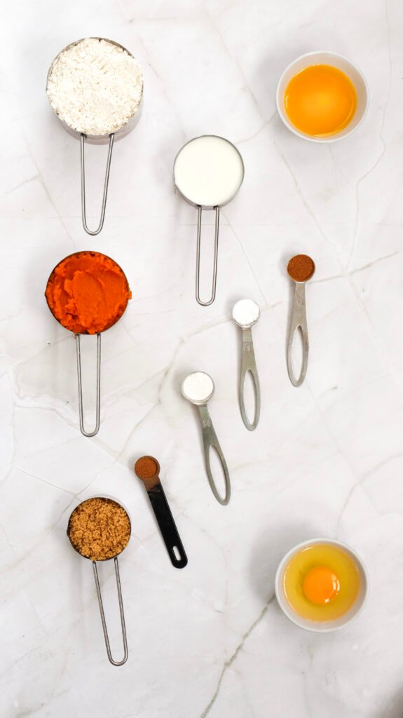 Pumpkin pancake ingredients, including flour, milk, eggs, pumpkin purée, brown sugar, melted butter, and spices in measuring cups and spoons