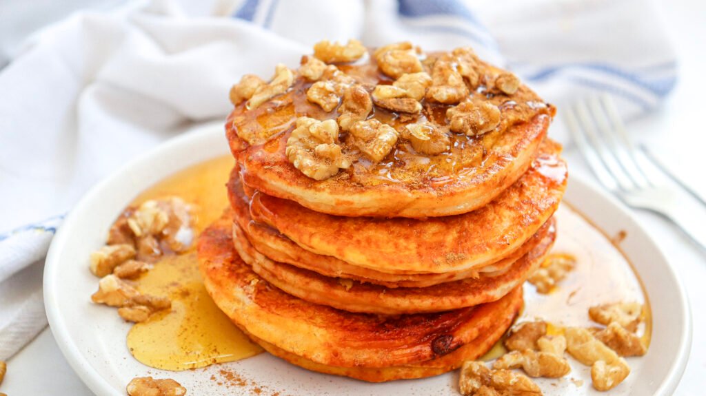 Stack of fluffy pumpkin pancakes topped with walnuts and maple syrup, served on a white plate