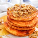 Stack of fluffy pumpkin pancakes topped with walnuts and maple syrup, served on a white plate