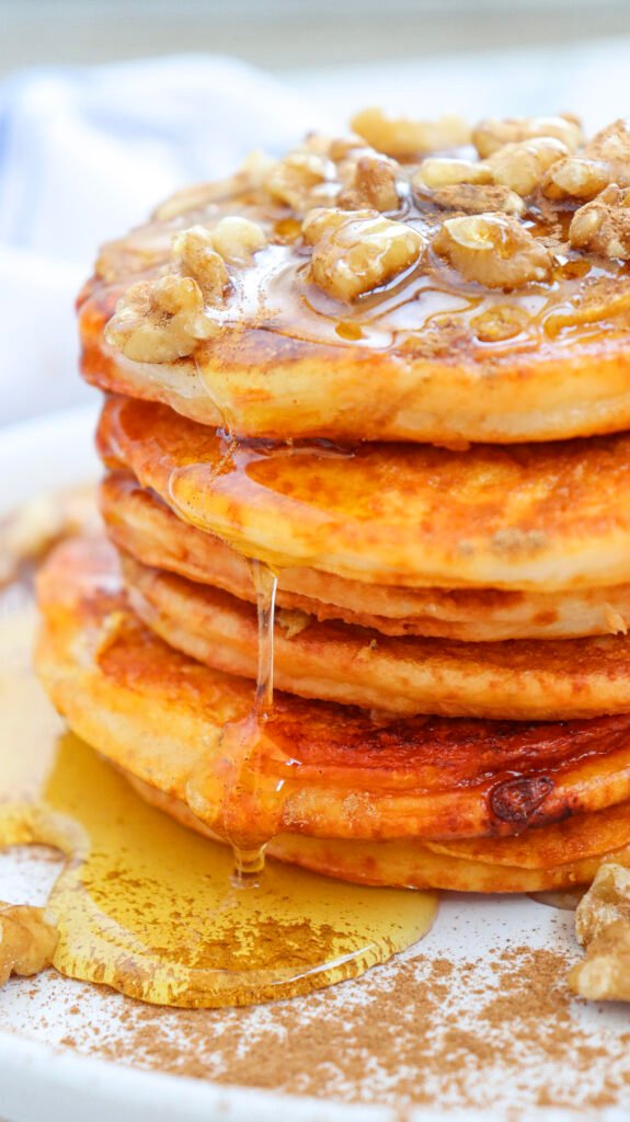 Stack of golden pumpkin pancakes topped with chopped walnuts and drizzled with maple syrup, with syrup dripping down the sides