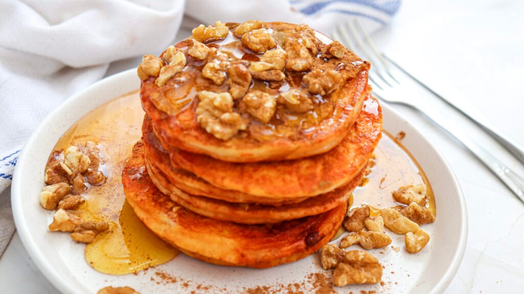 Stack of golden pumpkin pancakes topped with chopped walnuts and drizzled with maple syrup, with syrup dripping down the sides