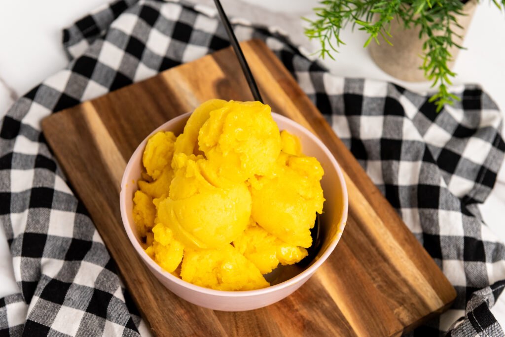 Scoops of vegan mango sorbet served in a pink bowl with a black spoon, set on a wooden board and black-and-white checkered cloth