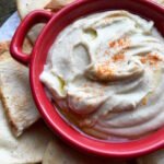 Creamy white bean dip in a red bowl, drizzled with olive oil and sprinkled with cayenne pepper