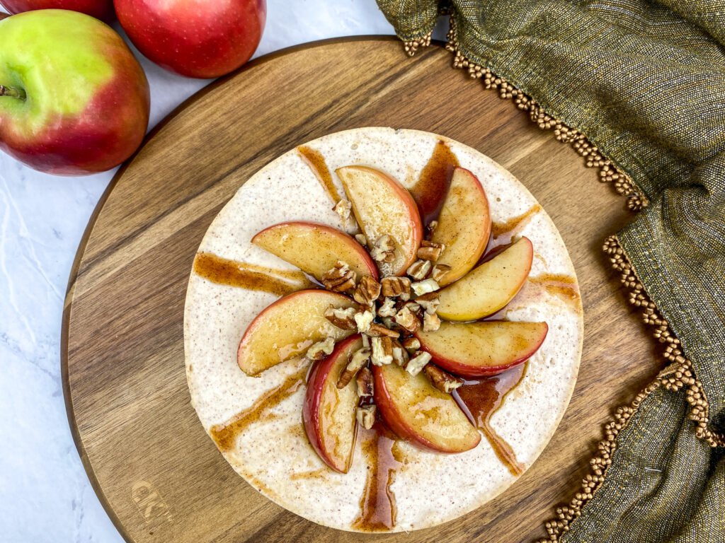 Overhead view of an apple cinnamon cheesecake topped with sliced apples, chopped pecans, and a drizzle of cinnamon sauce, served on a wooden board