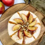 Overhead view of an apple cinnamon cheesecake topped with sliced apples, chopped pecans, and a drizzle of cinnamon sauce, served on a wooden board