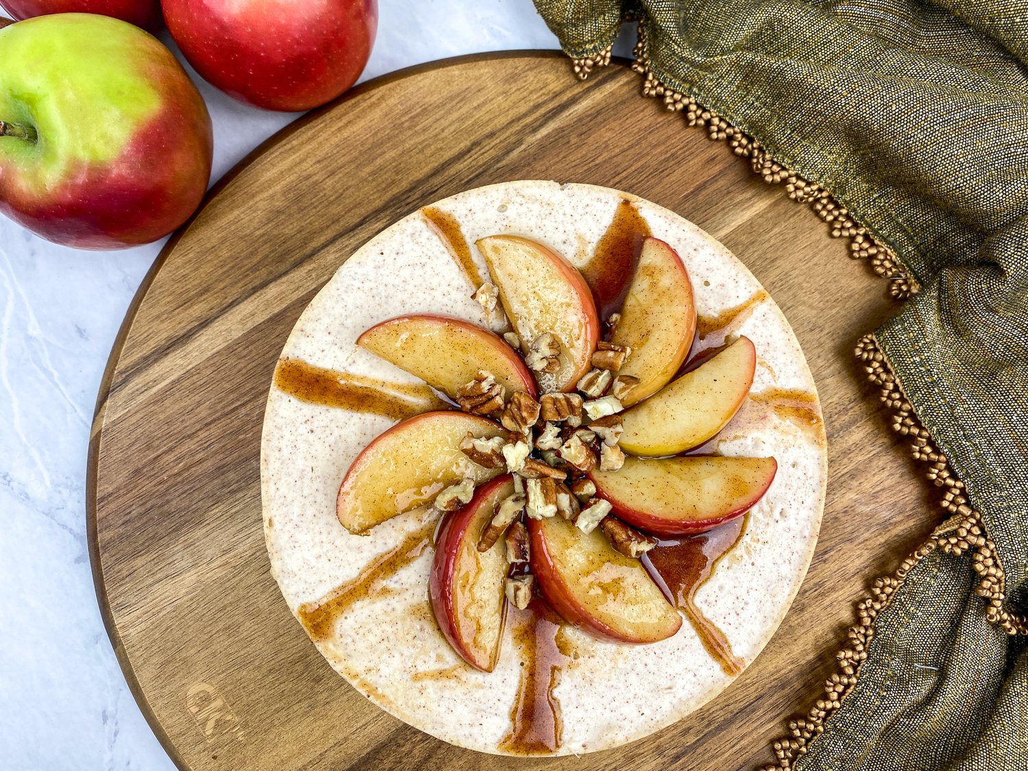 Overhead view of an apple cinnamon cheesecake topped with sliced apples, chopped pecans, and a drizzle of cinnamon sauce, served on a wooden board
