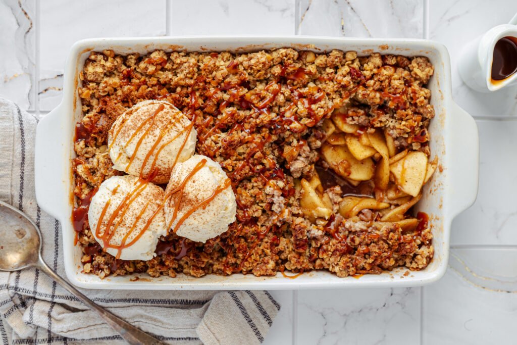 Baked apple crisp in a white dish topped with scoops of vanilla ice cream and drizzled with caramel sauce