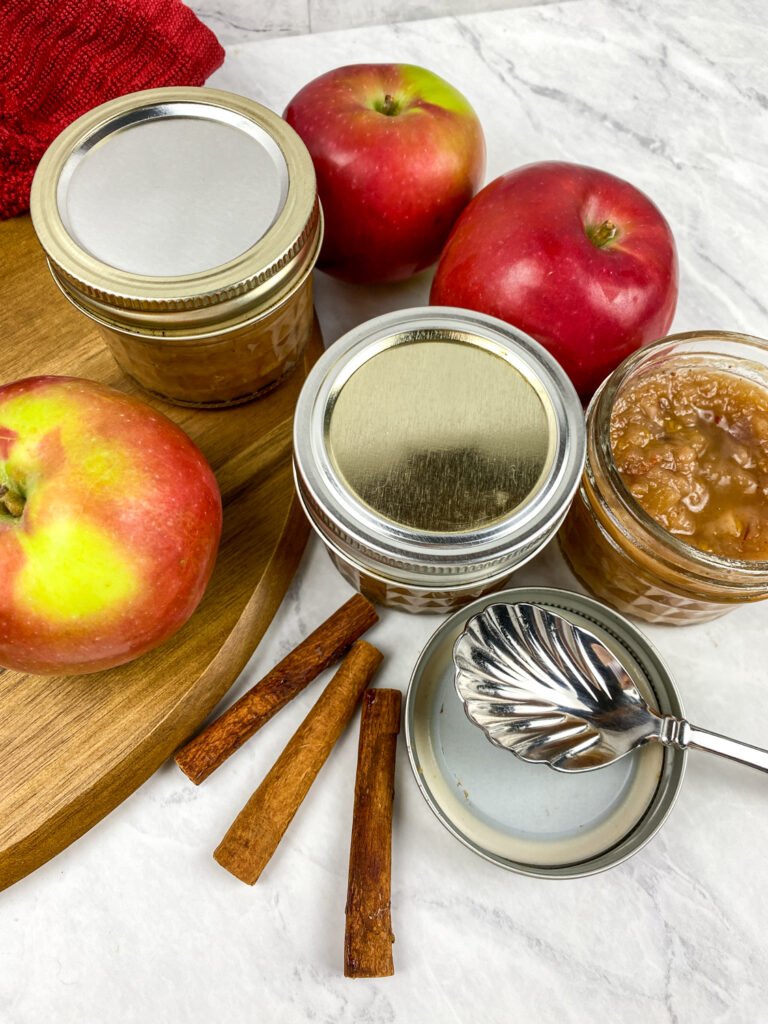 Jars of homemade savory apple chutney with fresh red apples, cinnamon sticks, and a decorative serving spoon on a white countertop