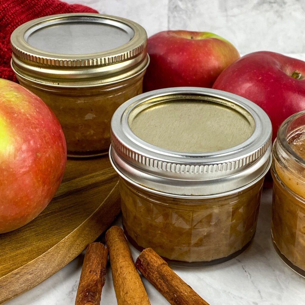 Homemade savory apple chutney in small glass jars, surrounded by fresh red apples and cinnamon sticks